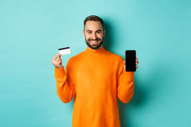 快乐网上购物快乐迷人的家伙展示手机屏幕和信用卡 微笑着满意 站在淡绿色的墙上信用时尚胡须