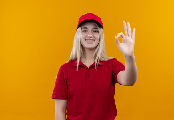 支撑微笑的小女孩穿着红色t恤 戴着帽子 戴着牙套 在孤立的橙色背景上显示出奥基的手势橙色牙科手势