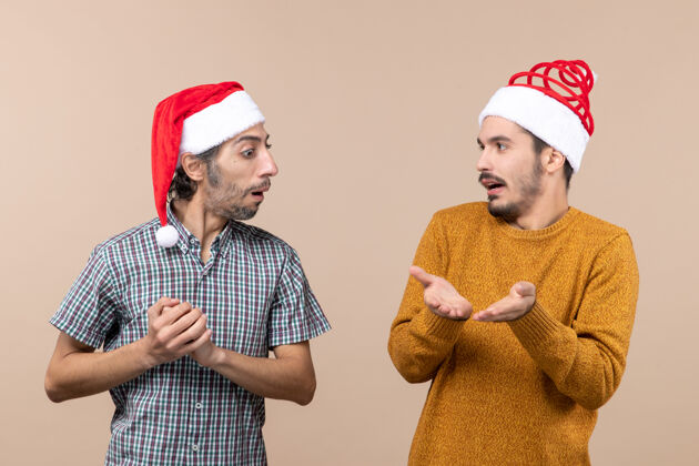 圣诞节前视图两个圣诞老人戴着圣诞帽 一个在米色背景上向另一个展示东西两个表演前面