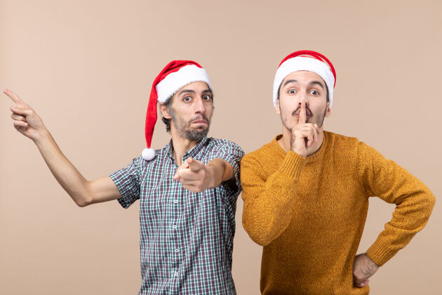 米色前视图两个戴着圣诞帽的家伙一个在展示什么 另一个在米色孤立背景上做嘘标志成人帽子视图