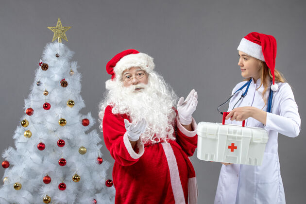 服装圣诞老人和女医生的前视图 女医生拿着急救箱在灰色的墙上人们前面圣诞老人