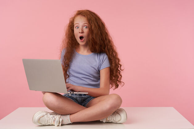 孩子美丽的红发女孩的肖像 长卷发盘腿坐在粉色背景下 穿着蓝色t恤和牛仔裤短裤 手持现代笔记本电脑 惊奇地看着相机发型肖像积极