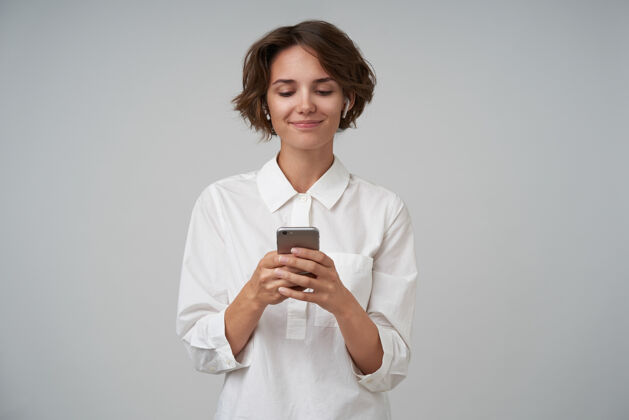 理发室内拍摄的一个可爱的年轻女子 棕色短发 穿着白衬衫 摆姿势 手里拿着智能手机 输入信息耳机表情肖像