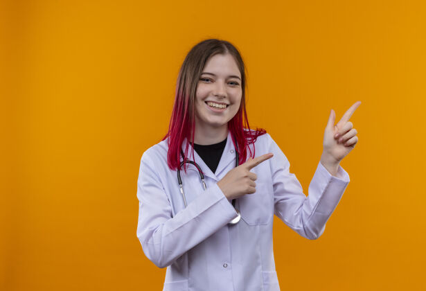 空间带着微笑的年轻医生女孩穿着听诊器医用长袍 手指指向一边 在孤立的橙色背景上留有复印空间年轻侧听诊器
