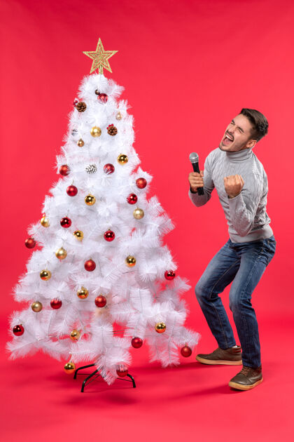 唱歌圣诞节的气氛与自信的家伙穿着牛仔裤站在装饰圣诞树附近 手持麦克风 唱他的歌自信圣诞自信的男孩