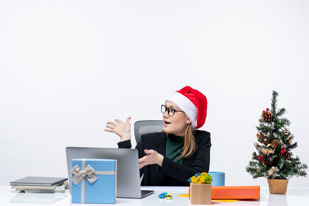 礼物新年气氛：年轻漂亮的女士戴着圣诞老人帽坐在桌子旁 桌上放着圣诞树和礼物 看着办公室里的东西圣诞节办公室圣诞老人