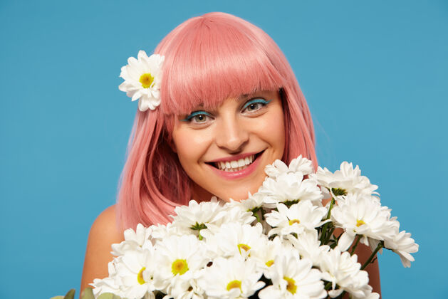 发型摄影棚的照片 年轻的浪漫的粉红色头发的女人与节日化妆摆在白色花朵蓝色背景 积极地看着相机迷人的微笑淑女洋甘菊20秒