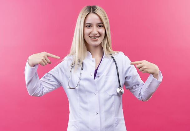 粉红色微笑的医生年轻的金发女孩穿着医用长袍戴着听诊器和牙套指着自己孤立的粉红色背景医生她自己医疗