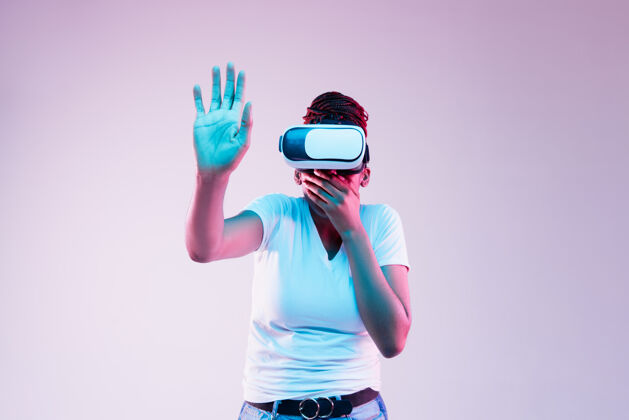 小玩意年轻的非洲裔美国女性在霓虹灯背景下 戴着vr眼镜在渐变背景下玩耍的肖像人类情感 面部表情 现代小玩意和技术的概念触动了某些东西虚拟现实虚拟现实
