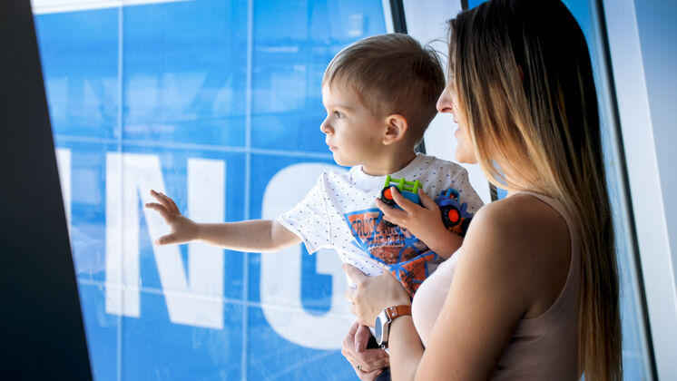 年輕年輕的母親抱著她的小兒子 透過機場候機樓的窗戶看的照片到達幼兒飛行