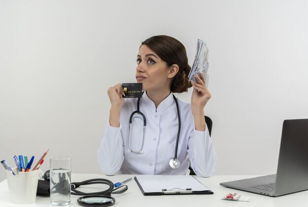 持有想着年轻的女医生穿着医用长袍 带听诊器 坐在办公桌旁 拿着医疗工具 拿着现金和信用卡 还有复印空间信用现金思维