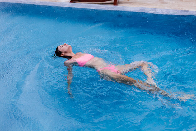 泳装在明亮的粉红色比基尼蓝色游泳池里的年轻快乐修长的欧洲女人游泳池夏天户外