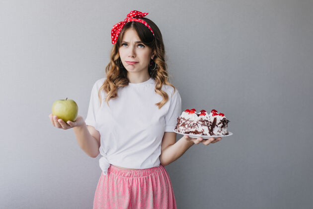 头发卷曲的高加索女孩在思考她的饮食室内拍摄的沉思的美妙女士拿着青苹果和奶油蛋糕漂亮蛋糕女性