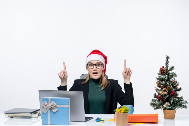 办公室好奇的女商人戴着圣诞老人的帽子坐在一张桌子旁 桌子上有一棵圣诞树和一份礼物 上面是白色的背景圣诞老人桌子商务