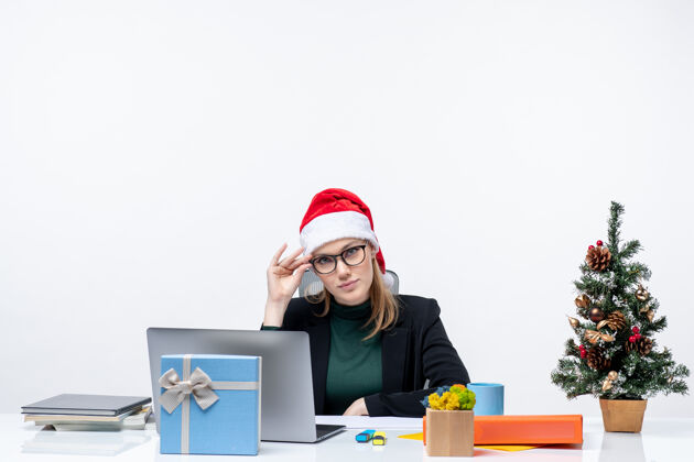 桌子新年气氛严肃迷人的女人 戴着圣诞老人帽坐在桌子旁 桌上放着圣诞树和白色背景的礼物人圣诞树圣诞老人