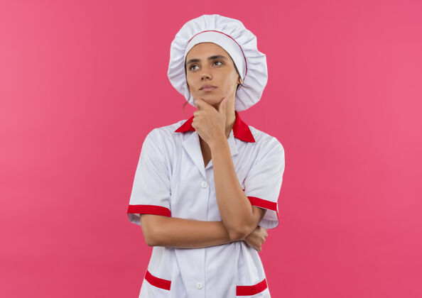 厨师看着一边想着年轻的女厨师穿着厨师制服把手放在下巴上孤立的粉红色墙壁与复制空间看侧穿
