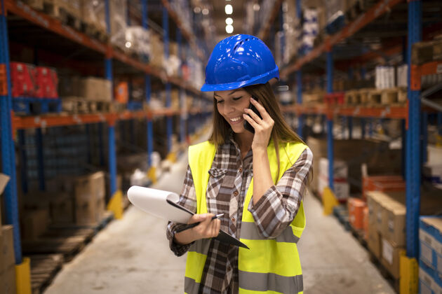 管理仓库工作人员戴着安全帽在电话上交谈 并在配送仓库设施中持有检查表供应物流质量