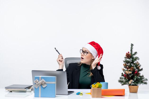 困惑一个困惑的金发女人 戴着圣诞老人的帽子 坐在一张桌子旁 桌子上有一棵圣诞树和一份白色背景的礼物圣诞节圣诞老人坐着
