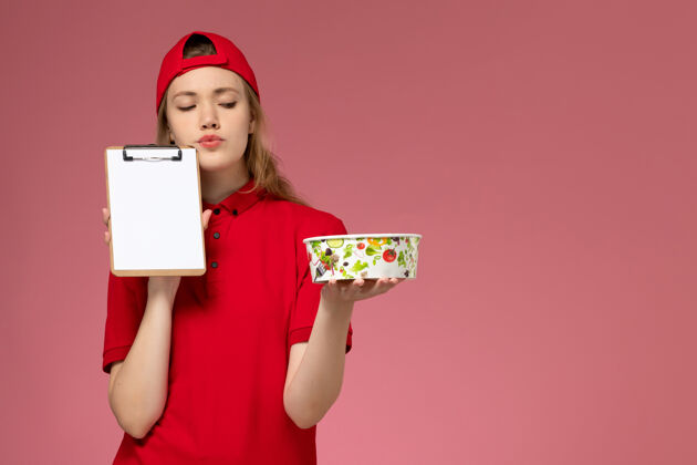 女人正面图身穿红色制服 披肩的年轻女快递员拿着投递碗和记事本在粉色墙上思考服务持有漂亮