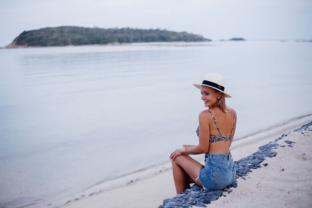 日落快乐的年轻欧洲女人穿着豹纹比基尼牛仔短裤和经典的白色帽子在热带异国情调的海滩上微笑着摆出有趣的俏皮女孩度假旅游的概念女人优雅穿着