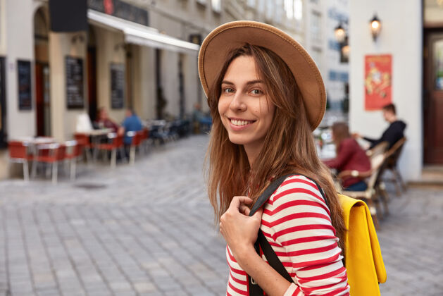 正面欢乐的欧洲游客漫步在城市街道的外景 背着背包 戴着棕色帽子 条纹毛衣城镇牙关高兴