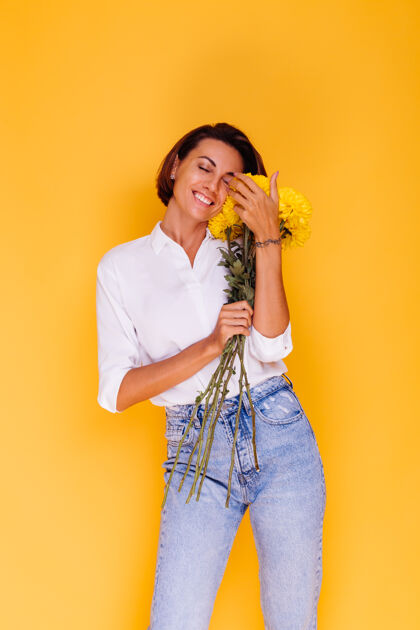 快乐摄影棚拍摄的黄色背景快乐的白人妇女短发穿着休闲服白衬衫和牛仔裤手持一束黄色紫苑微笑期待礼物