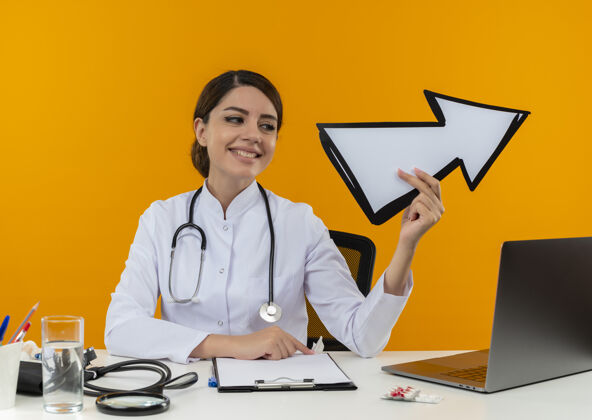 隔离看着面带微笑的年轻女医生 穿着医用长袍 手持听诊器 坐在办公桌旁 拿着医疗工具 拿着隔离黄墙上的方向标记 在电脑上工作听诊器电脑坐着