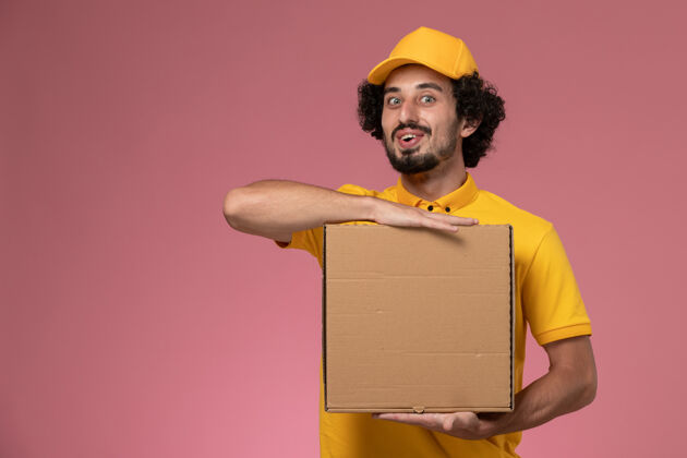 披萨盒子正面图：身穿黄色制服的男快递员拿着食品快递箱站在浅粉色的墙上拿着快递工作