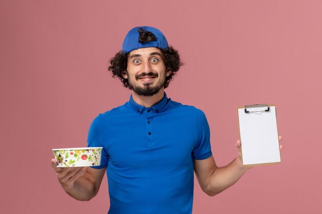 男性正面图：身穿蓝色制服 披风的男性信使拿着记事本和粉红色墙上的圆形投递碗碗成人正面