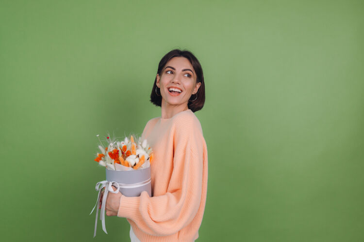 欢乐年轻女子穿着休闲桃色毛衣隔离在绿橄榄墙上手持橙白色花盒组成的棉花 吉普赛拉小麦和拉古鲁斯作为礼物开心惊喜积极欢呼花