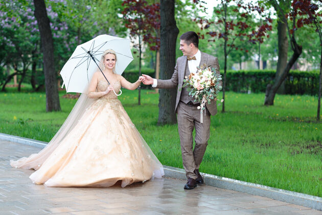 女人快乐的新郎新娘打着白色雨伞 在夏天的公园里露天婚礼毕业白天雨伞