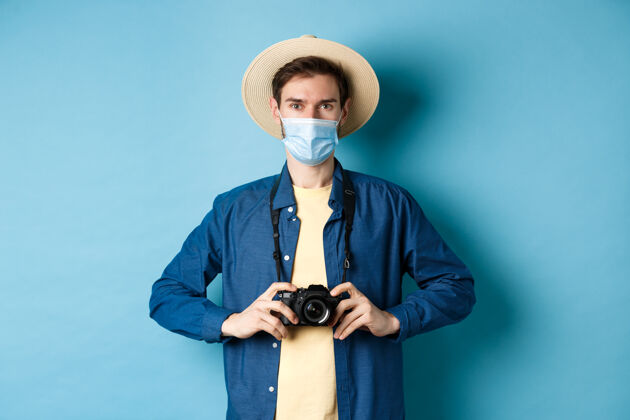 胡须Covid-19 流行病和旅行概念年轻人带着相机出国旅行 度假时拍照 戴着冠状病毒的医学面具 蓝色背景冠状病毒肖像人