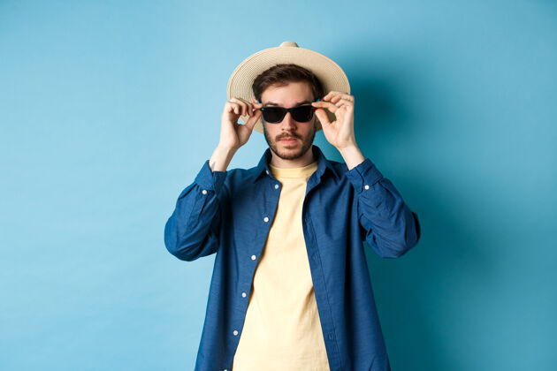 太阳镜穿着草帽的帅哥游客放暑假戴上墨镜 站在蓝色的背景上帅哥年轻脸