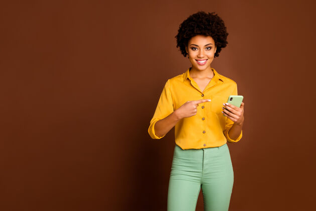 情绪正面开朗的黑人女学生肖像使用智能手机搜索网上销售指示点食指装置穿时尚黄色裤子隔离棕色微笑女人美国