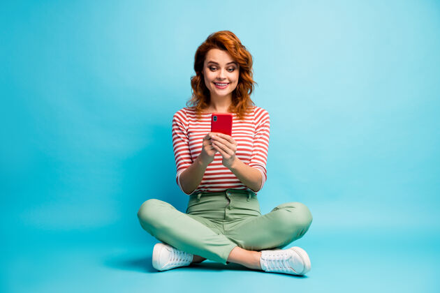 女性正面开朗女人的全身照片双腿交叉坐着使用智能手机阅读分享社交媒体新闻穿着时尚的衣服蓝色隔离女孩交叉互联网