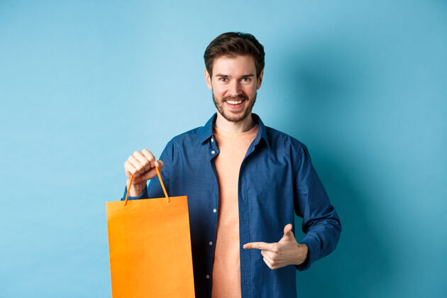 人站在蓝色背景上 微笑着的白人男子用手指指着装有购买物品的橙色购物袋包朋友年轻