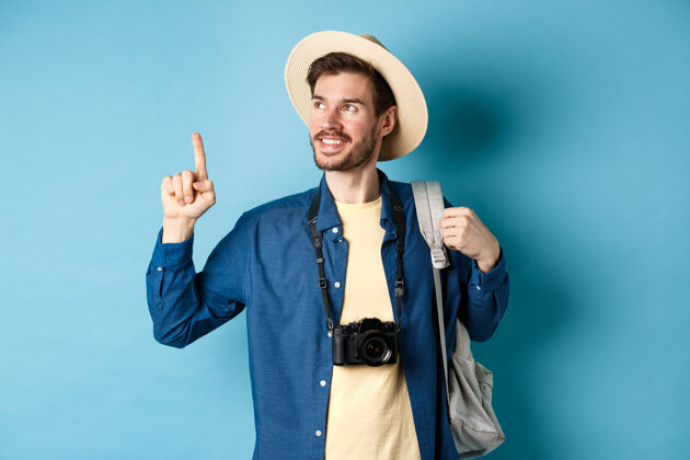帽子度假时帅气的帅哥 戴着夏帽 抬头看着logo 高兴地笑着 拿着相机和背包站在蓝色的背景上年轻休闲指着
