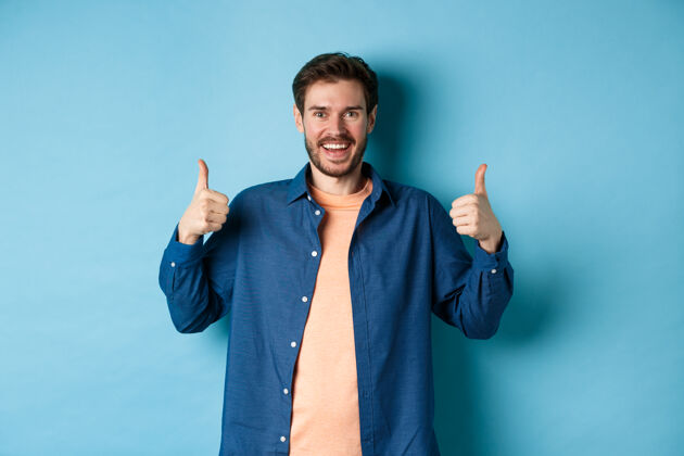 随意一个穿着休闲衬衫的高加索人站在蓝色的背景上 兴高采烈地竖起大拇指表示赞同 微笑着称赞一些好东西站立脸推荐