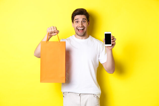 应用程序惊讶的男人拿着购物袋 展示智能手机屏幕模特礼品休闲