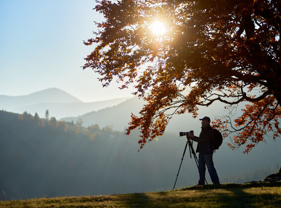 远足徒步旅行者带着相机在大树下的山景背景下的草谷里全景独自草地