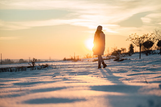 阳光一个穿着暖和衣服的女人独自站在雪白的草地上 欣赏冬日乡间如画的日落无法辨认女人日落