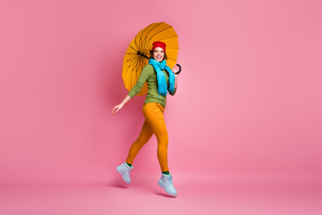 积极开朗甜美可爱女孩的全尺寸照片跳跃享受散步抱着明亮的阳伞戴着蓝红色的头饰毛衣裤子鞋子隔着粉色的墙去围巾休闲