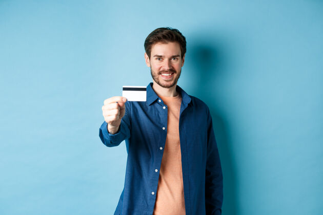 肖像开朗的白人男子 微笑着出示塑料信用卡 推荐银行 站在蓝色背景上情感脸办公室