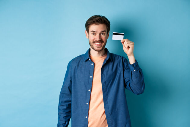 人快乐的白种人微笑着 展示着塑料信用卡 穿着蓝色背景的休闲服站着年轻肖像成人