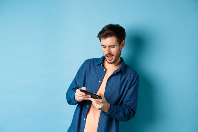 成人年轻人用手机玩电子游戏 身体倾斜 手持智能手机水平 站在蓝色背景上发型男手机