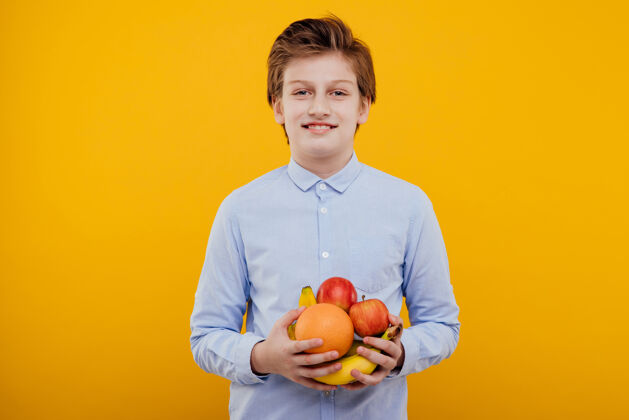 青少年手里拿着水果 穿着蓝色衬衫 孤零零地站在黄色的墙上的漂亮的未成年男孩小学生颜色男孩