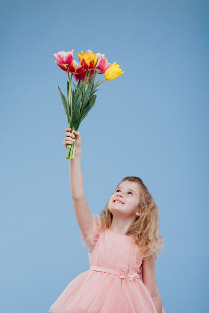 裙子郁金香微笑的小女孩拿起一束春天的花朵 粉红色的裙子 孤立在蓝色的墙上童年快乐看