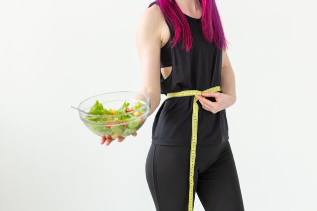 强壮一位紫色头发 身份不明的年轻女运动员测量着卷尺的腰 手里拿着一份蔬菜沙拉健身房健康锻炼