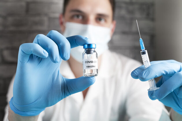 护理研究人员手里拿着Covid-19疫苗 男医生穿着白大褂 戴着医用口罩 戴着蓝色手套 手里拿着装有治疗冠状病毒疫苗的注射器和瓶子冠状病毒治疗的概念外科手术Sars测试