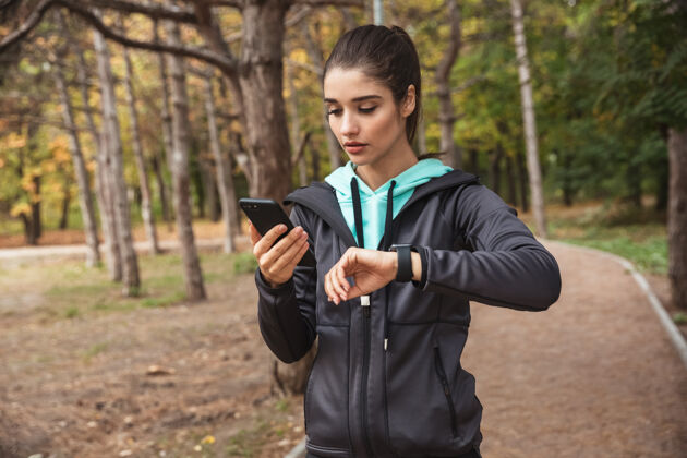 年轻令人惊叹的年轻漂亮的健身女士在公园户外用手机看手表的照片放松耳机户外